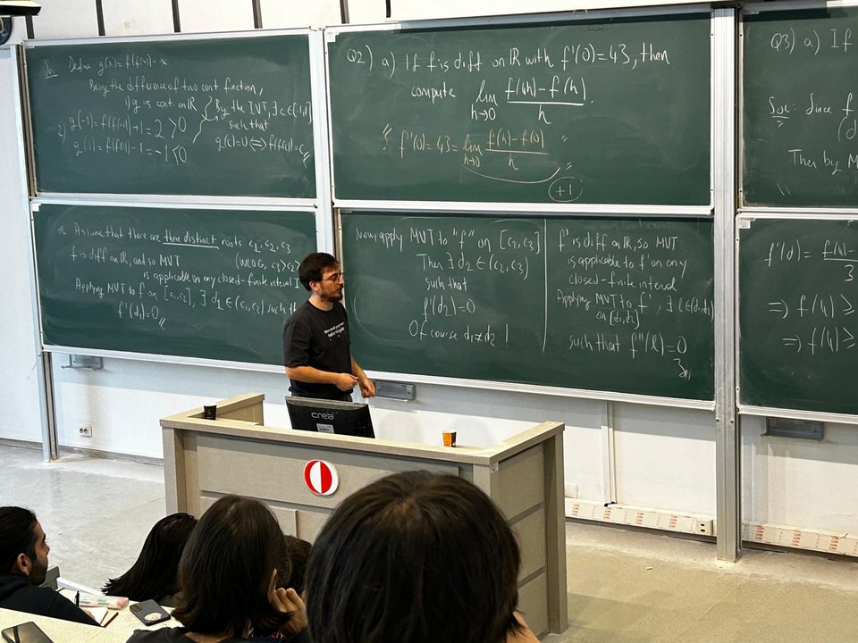 A lecture theatre at ODTU with three blackboards covered in handwritten analysis proofs, the lecturer at the podium.