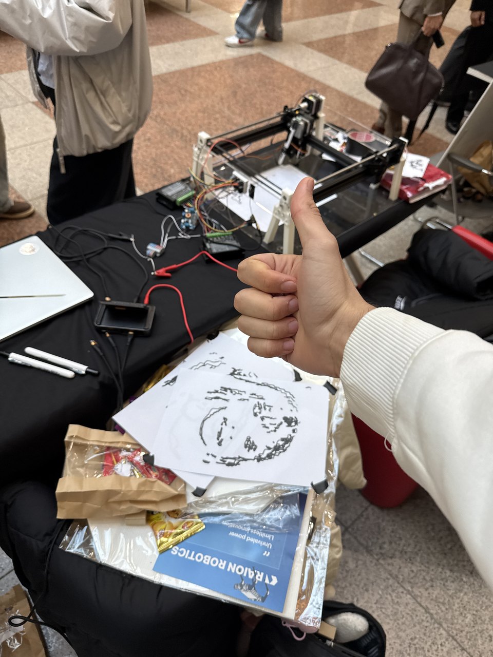 The drawing machine on a table with a finished line-drawn portrait beside it, and a hand giving a thumbs up.