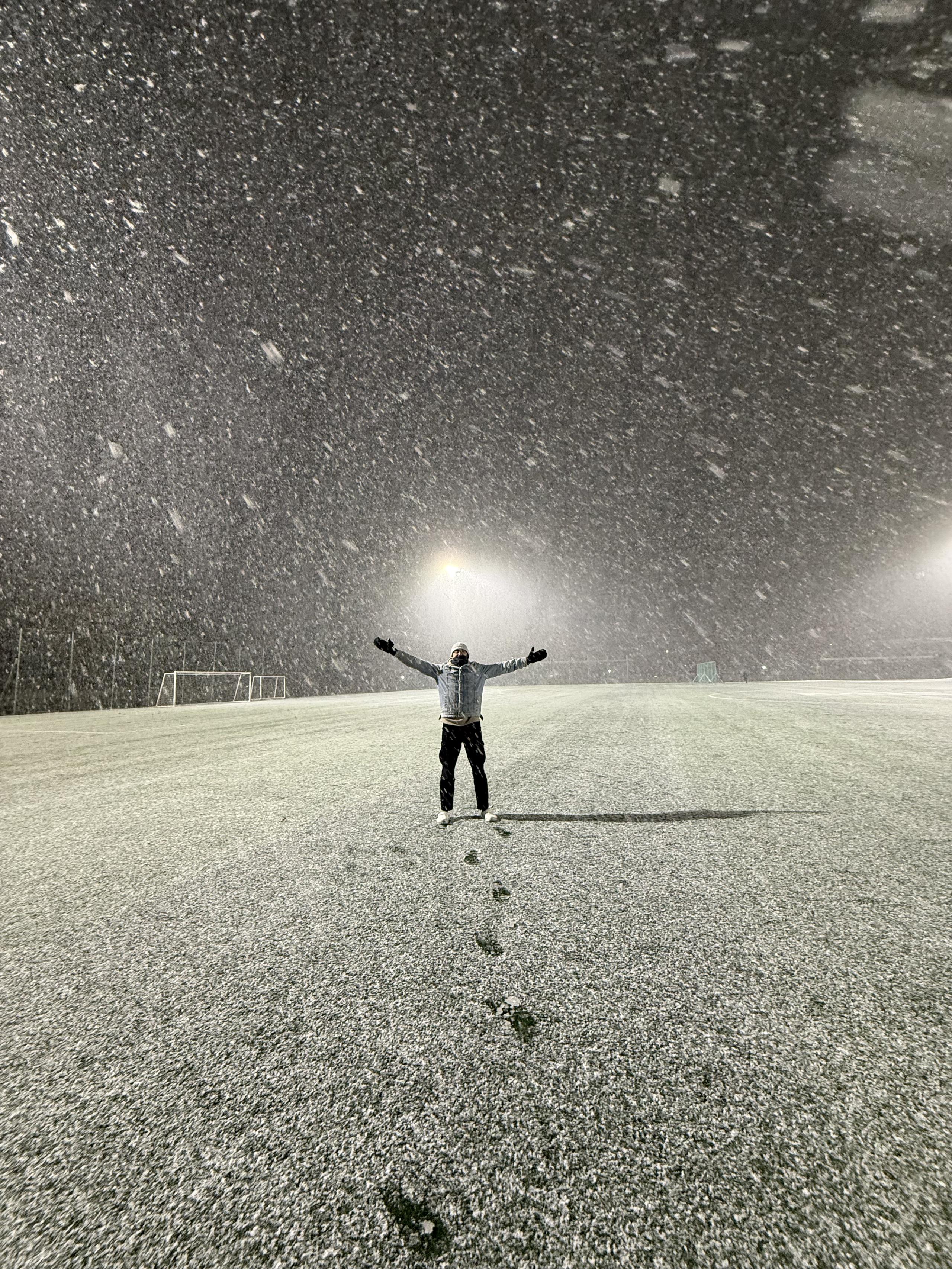 Abdullah alone on a floodlit sports field in heavy snow at night, arms spread wide, his footprints leading back across the pitch.