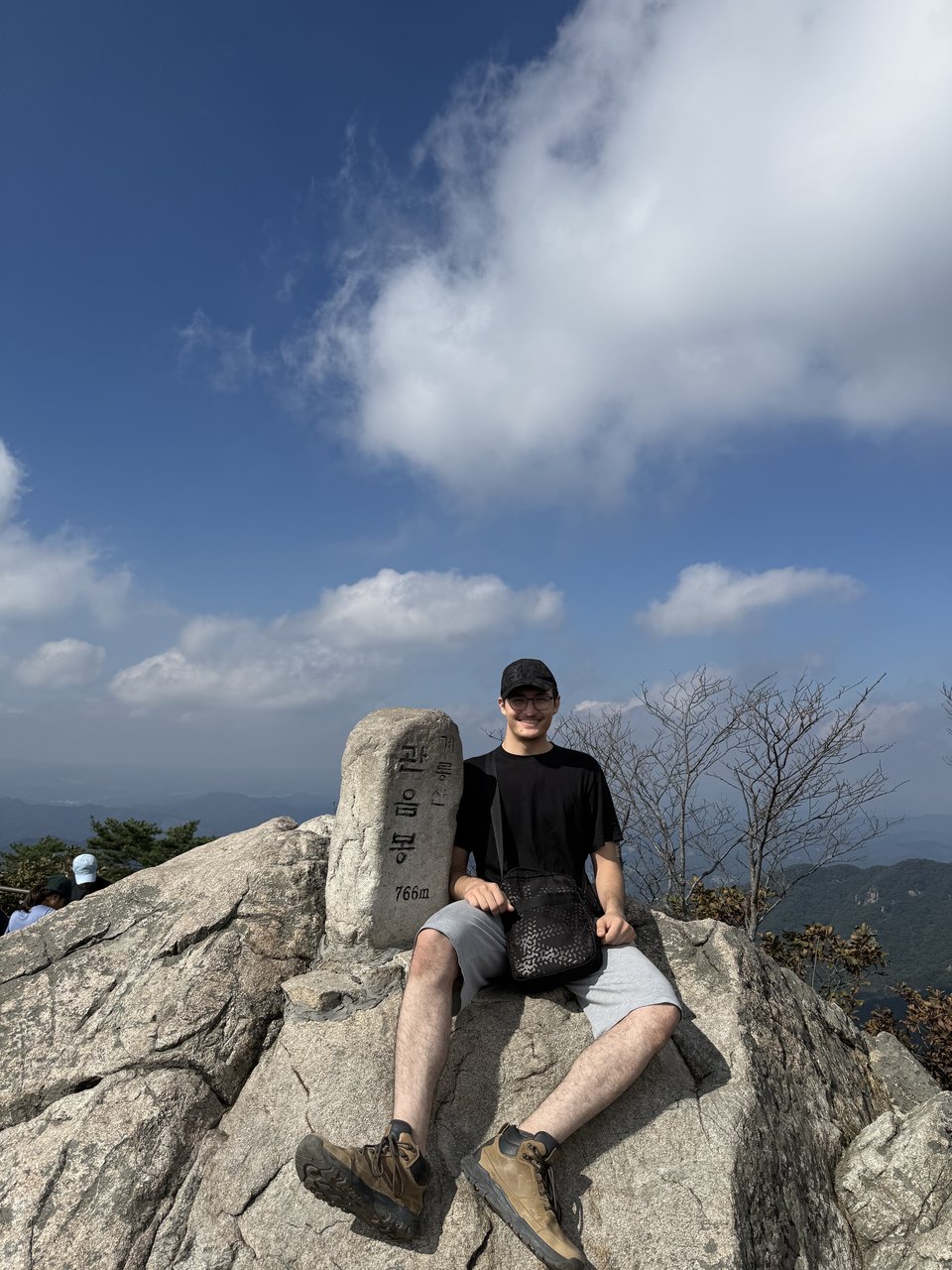 Abdullah sitting on a rock at a mountain summit beside a stone marker, deep blue sky behind him.