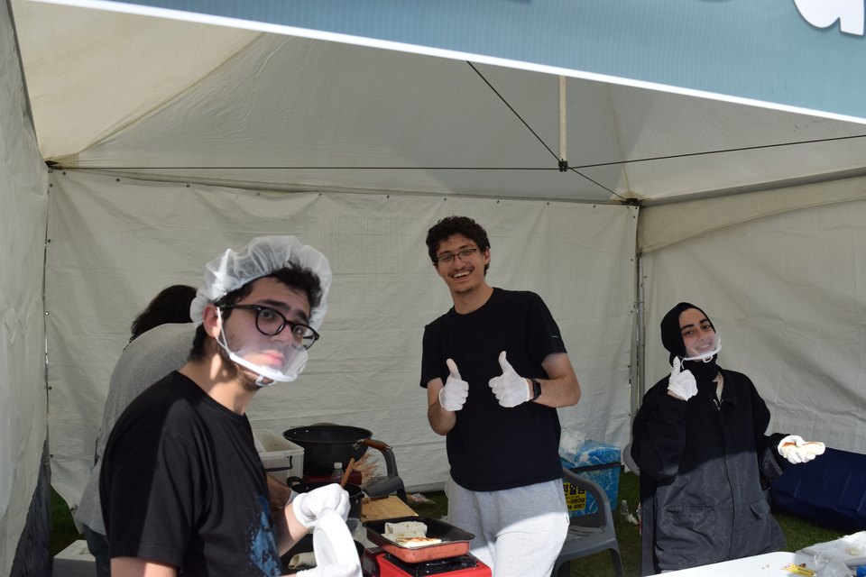 Abdullah and two friends in hairnets and gloves behind a food stall under a marquee.