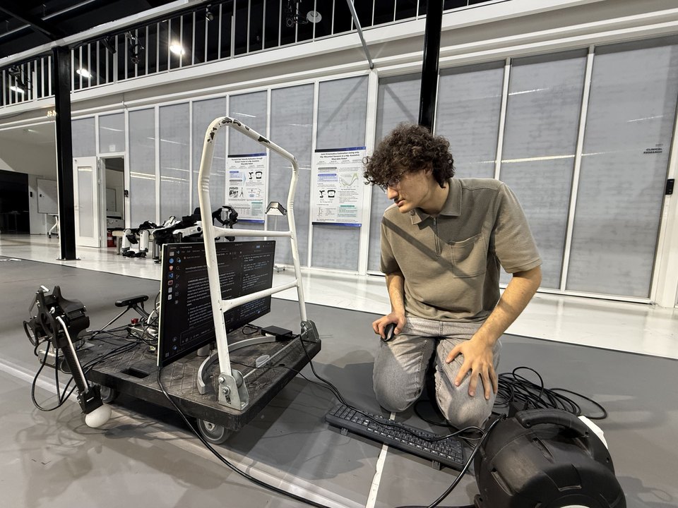 Kneeling beside the dual-arm cart on the hackathon floor: two arms bolted to a flatbed trolley, a monitor and keyboard on the deck.