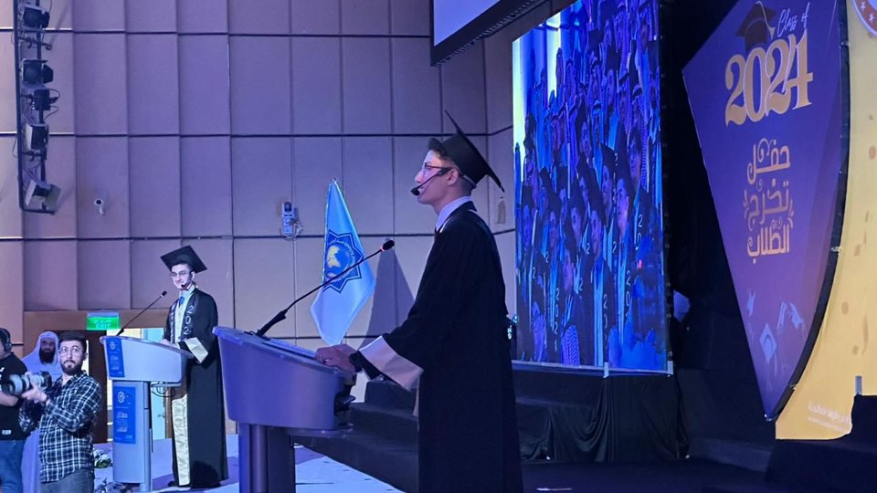 Abdullah at a lectern on a blue-lit stage in front of a large 2024 backdrop, addressing the graduation audience.