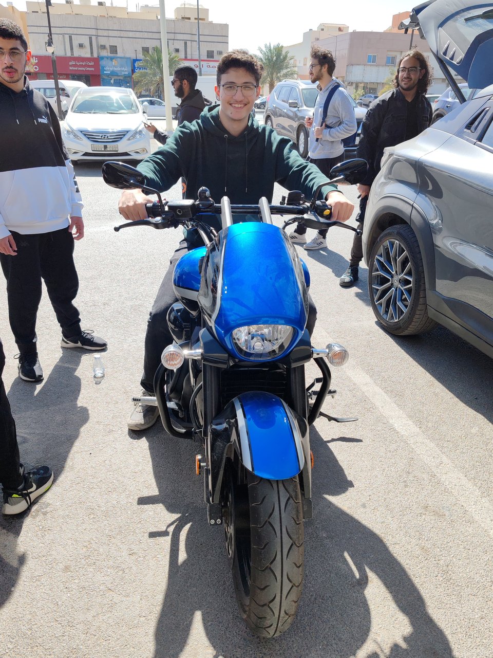 Abdullah sitting on a blue motorbike in a sunlit street with friends standing around.
