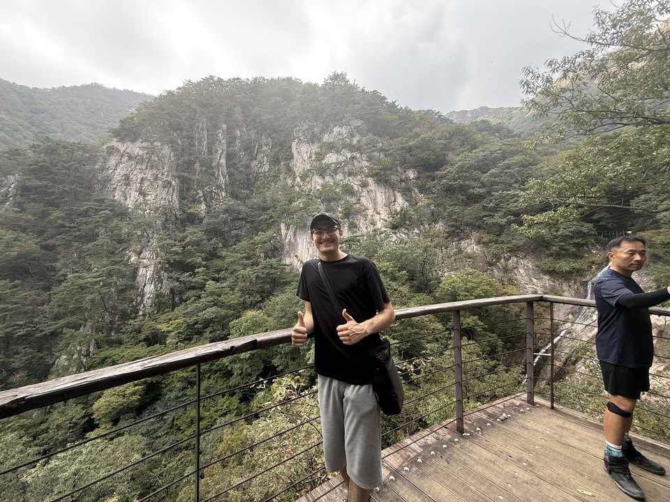 Abdullah on a mountain boardwalk with a cliff face and forest behind him.