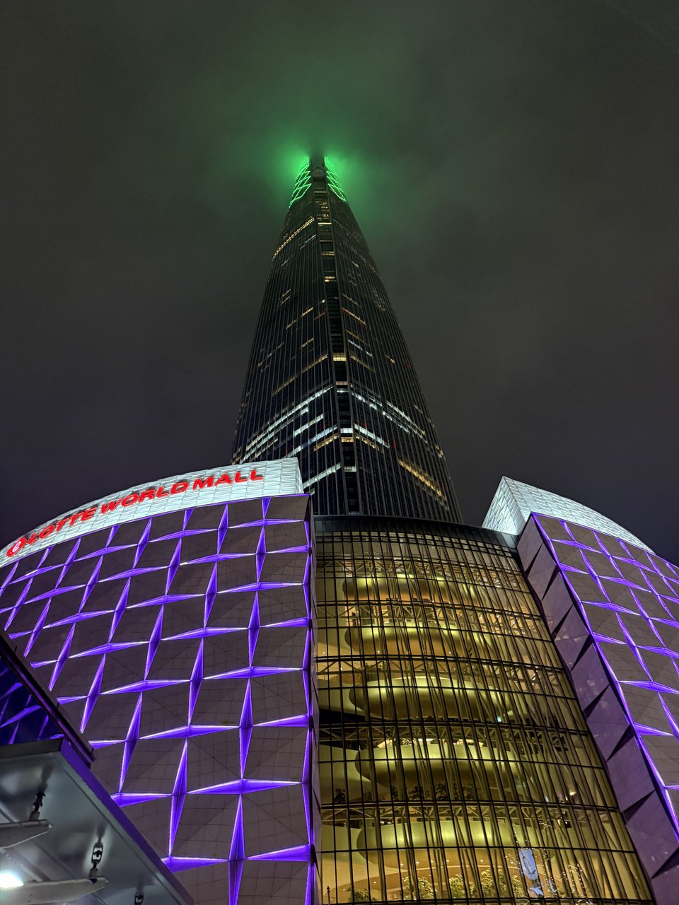 A tall tower lit green at night above a glowing shopping complex.
